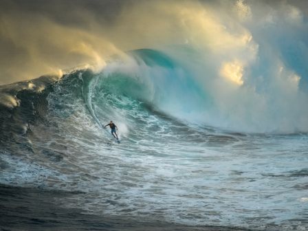 A lone surfer rides a towering wave under dramatic, sunlit clouds, carving through a turquoise barrel with spray and mist all around.