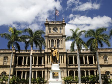 A historic building with a central clock tower, statue in front, and palm trees lining the facade. A flag waves atop the tower.