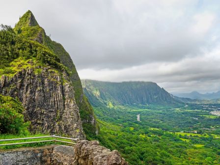 A rugged cliff with a sharp peak overlooks a lush, green valley and distant hills under a cloudy sky.