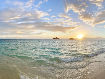 A serene beach at sunset with gentle waves, turquoise water, and a couple of small rocks on the horizon against a partly cloudy sky.