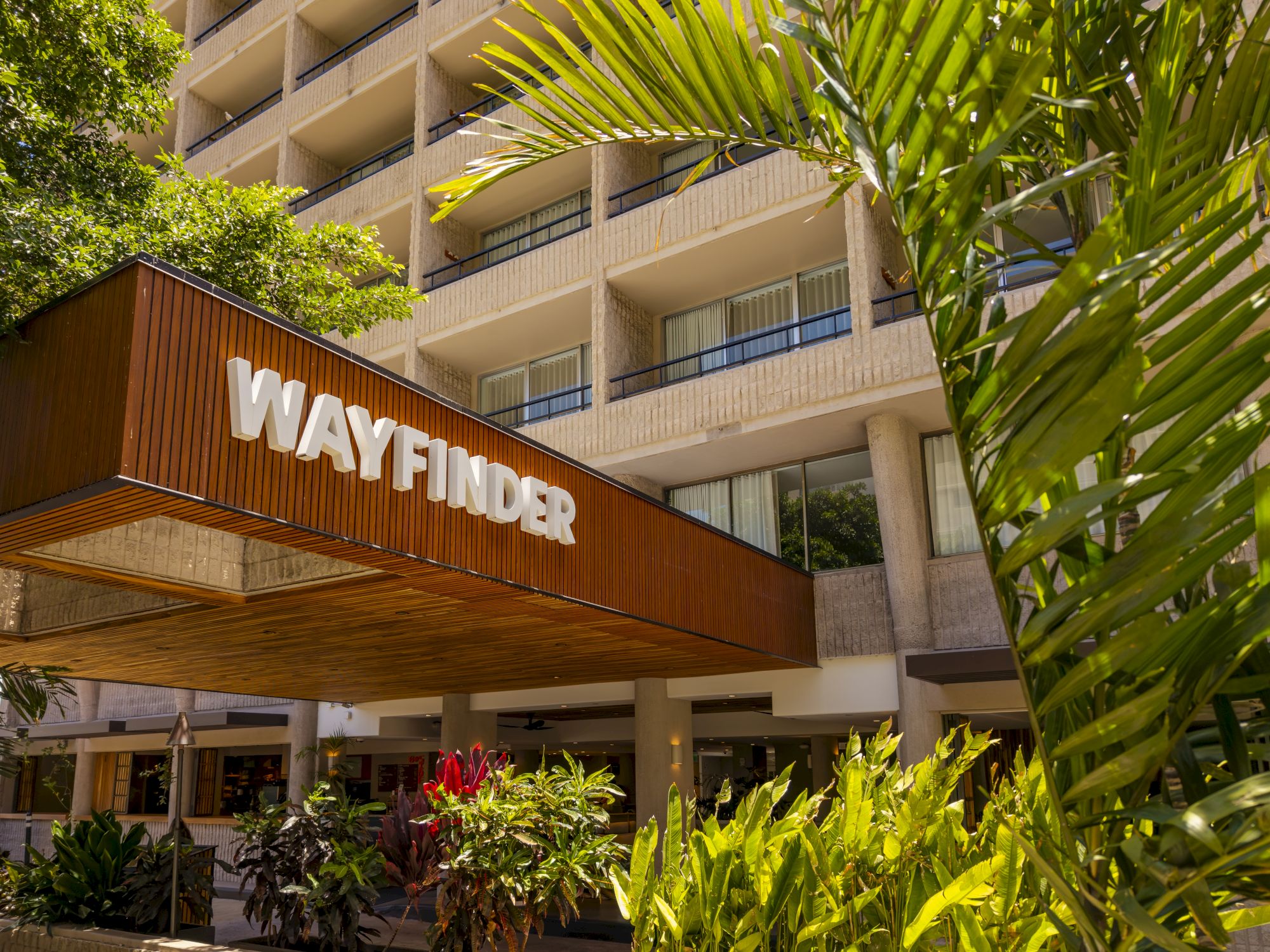 A Wayfinder sign on the entrance of a tropical, multi-story building with lush plants and palm fronds, likely a hotel or resort.