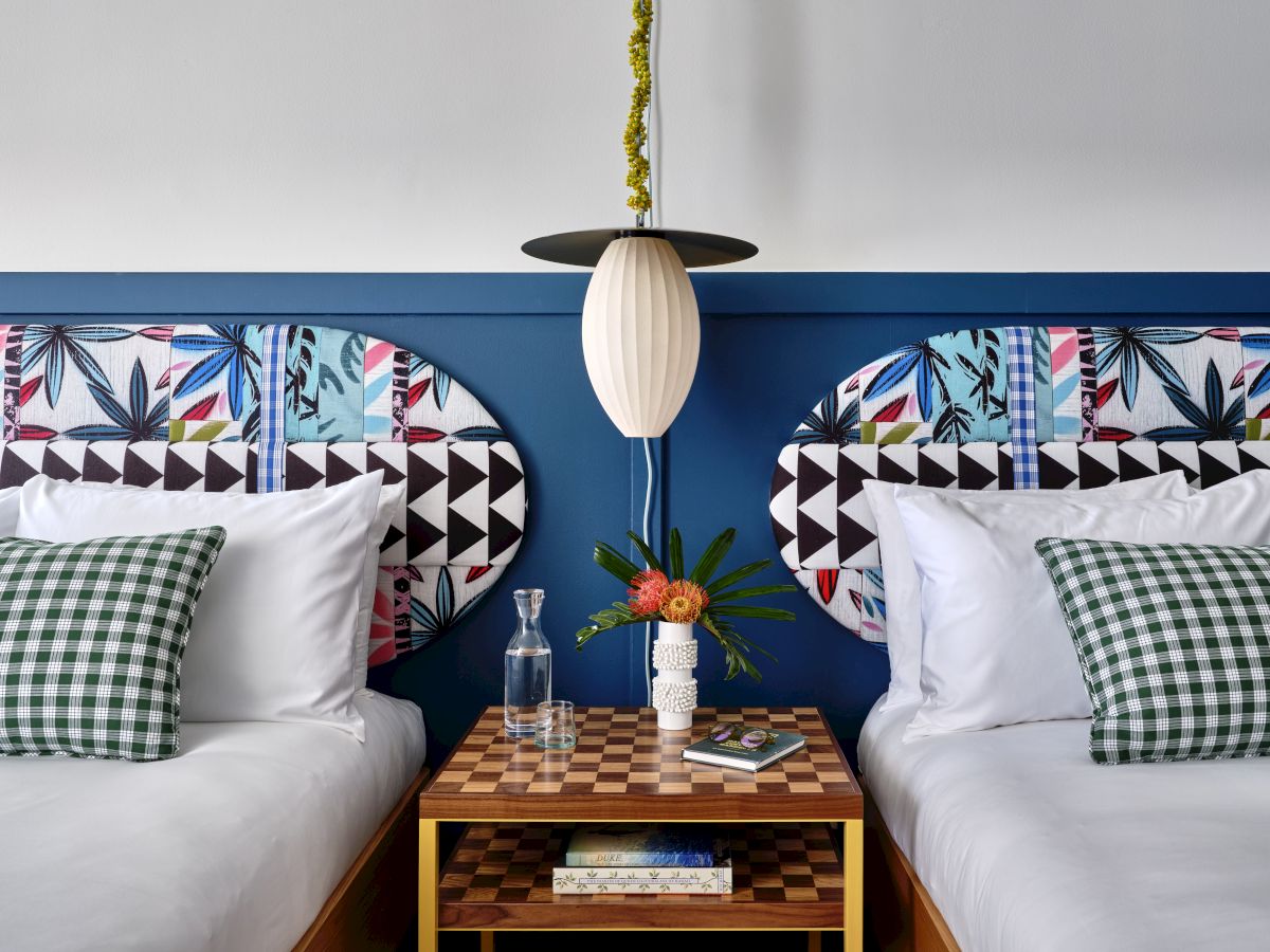 Two twin beds with colorful, geometric headboards meet in a modern hotel room; a shared nightstand holds a bottle, flowers, and a book, under a hanging lamp.