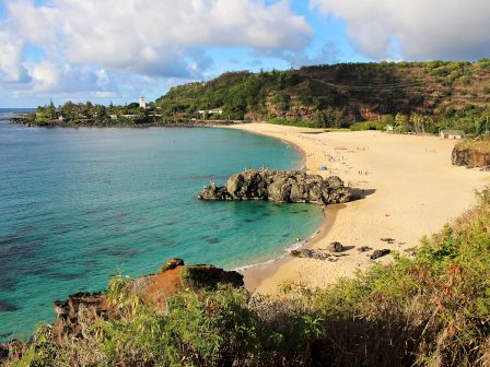 A tropical beach with turquoise water, golden sand, rocky outcrops, and lush green hills under a partly cloudy sky, paradise by the sea.