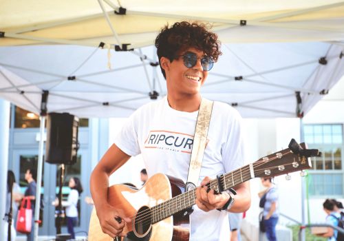 A smiling young man with curly hair wearing sunglasses plays an acoustic guitar under a white canopy at an outdoor event.