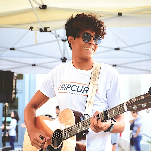 A smiling young man with curly hair wearing sunglasses plays an acoustic guitar under a white canopy at an outdoor event.