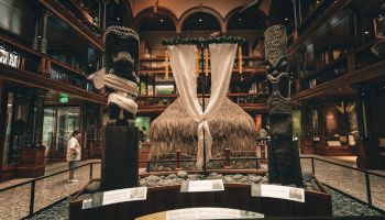 A grand, dimly lit museum hall with a central thatched hut exhibit, dark wood balconies, hanging drums, and display plaques around a circular platform.
