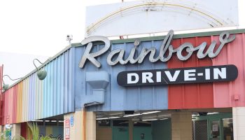 Rainbow Drive-In storefront with retro signage, blue and pink facade, outdoor seating, and a trash can outside.