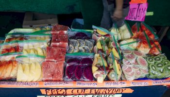 A colorful fruit stand with sliced fruit in clear rows; signs read &ldquo;Fresh Cut Everyday&rdquo; and flavors like mango, watermelon, dragon fruit, and kiwi.