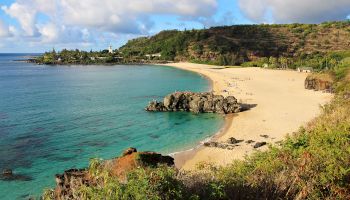 A tropical beach with golden sand, turquoise water, rocky outcrops, and green hills in the background, under a partly cloudy sky.