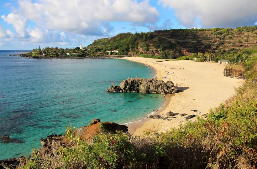 A tropical beach with golden sand, turquoise water, rocky outcrops, and green hills in the background, under a partly cloudy sky.