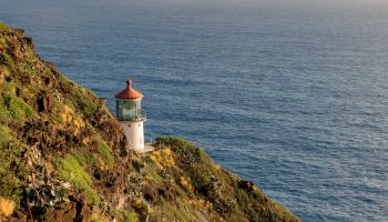 A lighthouse perched on a rocky cliff overlooking a vast blue ocean under a partly cloudy sky, with warm sunlight on rugged terrain.