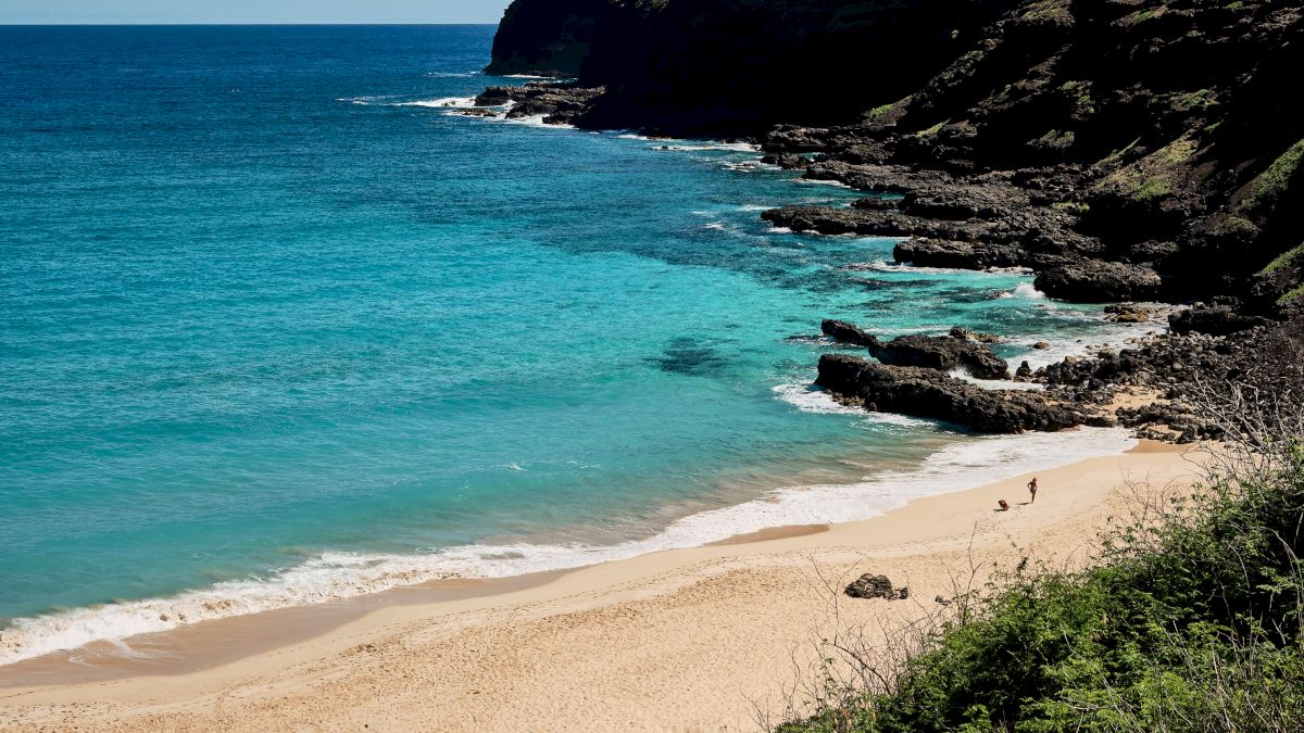 A sunny beach with turquoise water, a sandy shore, and a rocky green hillside jutting into the blue ocean, under a clear sky.