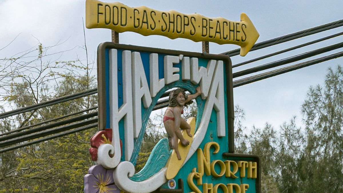 A weathered sign for &ldquo;LANI North Shore Shop&rdquo; with a shark mascot and a rooster, advertising food, gas, and shops along a highway.