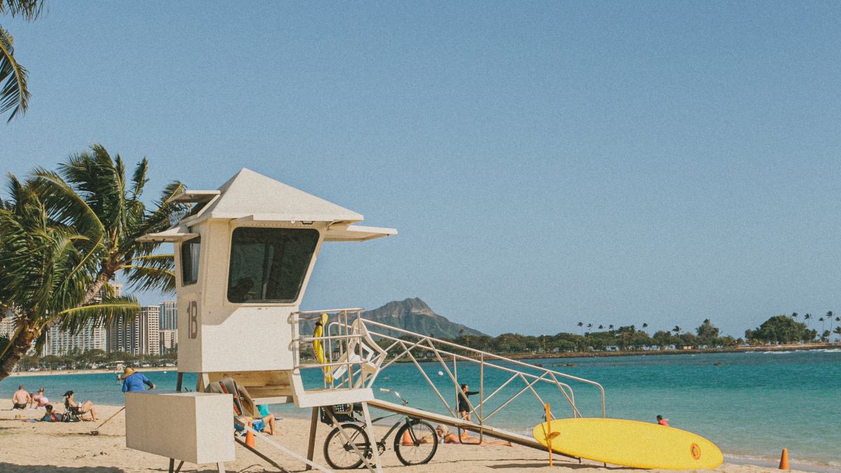 A lifeguard tower stands on a sunny beach with a yellow kayak nearby, palm trees in the background, and orange cones dotting the sandy shore.