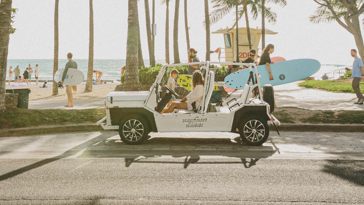 People enjoy a tropical beach scene with a white SUV parked on the road, tall palm trees, and surfers by the shore, sunny and relaxed.