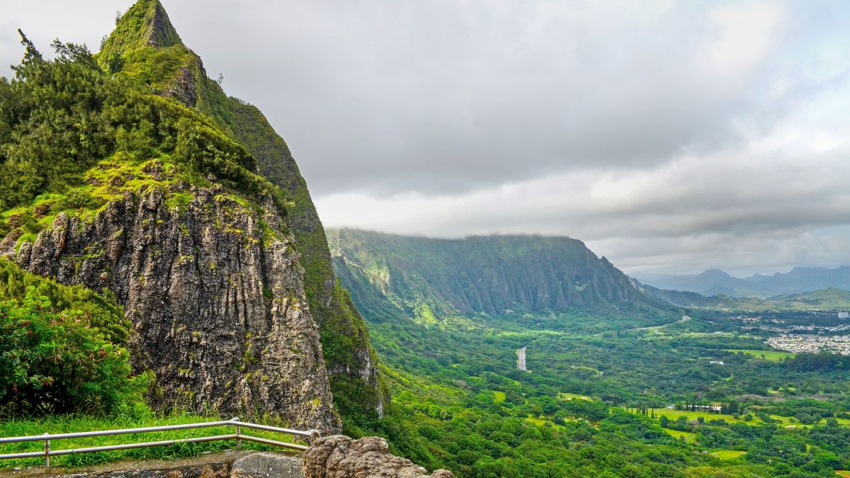 A lush, rugged mountainside with a sharp peak, green cliffs, and a valley below dotted with fields and distant hills, under a cloudy sky.