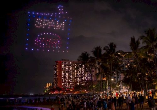 A crowded beach at night with tall palm trees, a lit cityscape, and a glowing fair-like square of lights in the sky above the crowd.