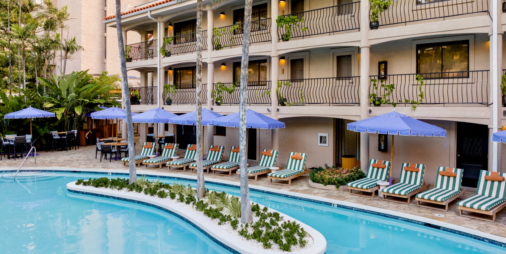 A resort pool area with clear blue water, palm trees, striped lounge chairs, blue umbrellas, and a beige hotel building with balconies surrounding the pool.