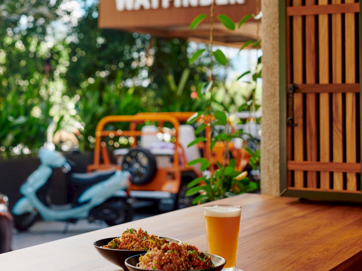 A cozy cafe scene with a wooden counter, bowls of food and a beer, outdoors sign reading &ldquo;Wayfinder&rdquo; and greenery in the background.
