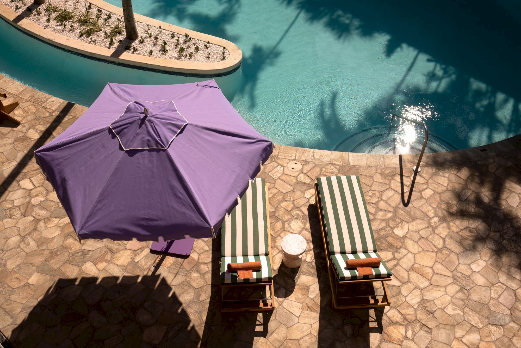 A sunny poolside with two striped lounge chairs under a purple umbrella beside a turquoise pool, casting shadows on a stone patio.