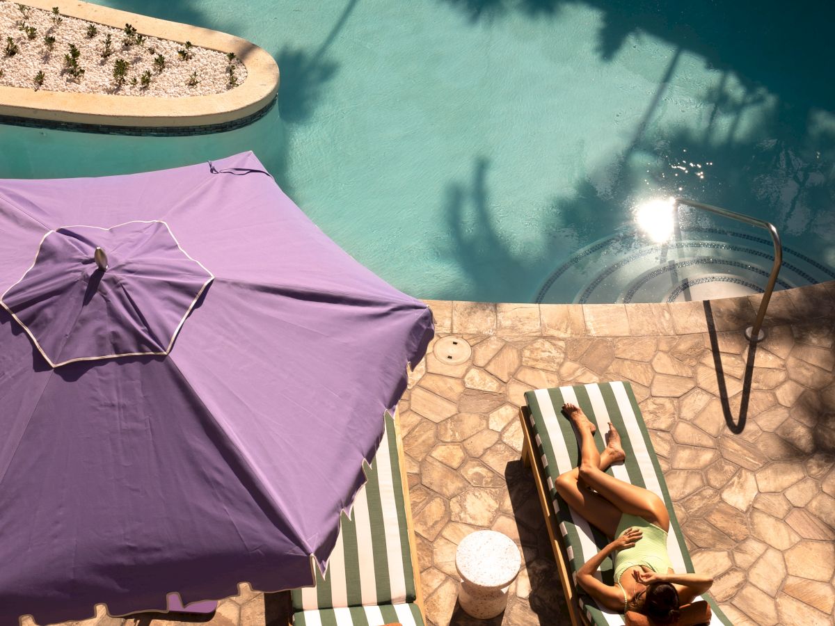 Two people relax on lounge chairs by a turquoise pool under a purple umbrella at a sunny resort.
