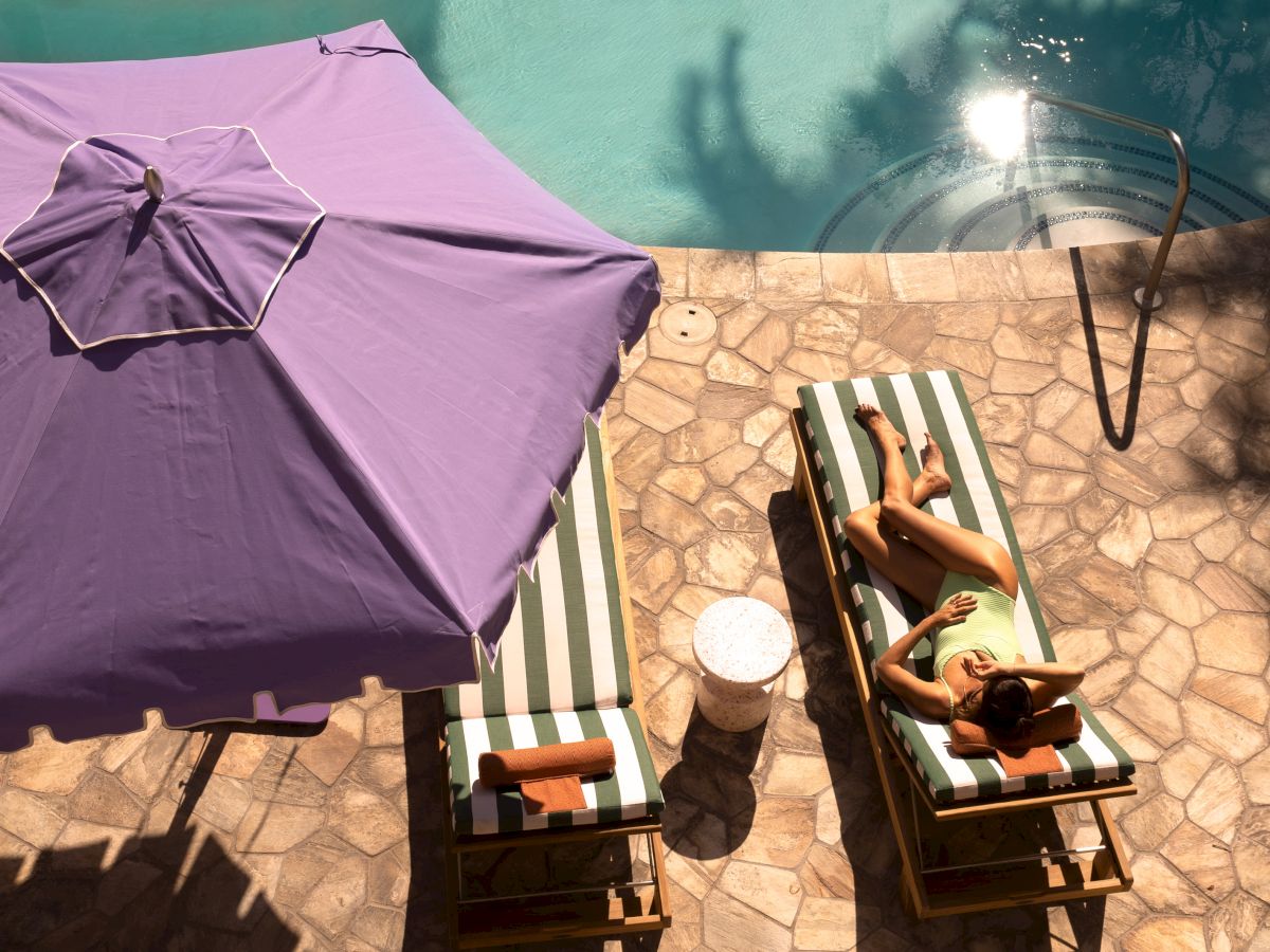Two people sunbathe under a purple umbrella by a pool, with a striped chair setup, a towel, and shadows on a stone pool deck.