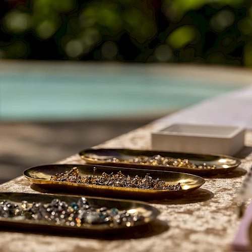 A row of shiny, bejeweled bracelets laid out on a sunlit outdoor surface near a blurred pool, catching glints of light as they sparkle.