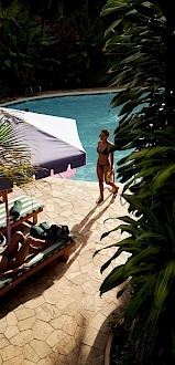People relax by a pool area with sun loungers, an umbrella, and lush tropical plants surrounding the stone patio.