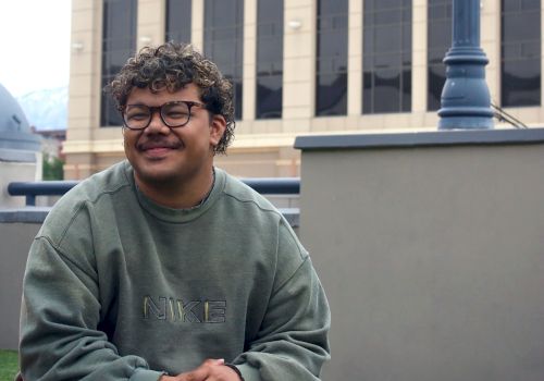 A young man with curly hair and glasses sits on grass near a modern beige building and lamp post, wearing a green sweatshirt and shorts, smiling.