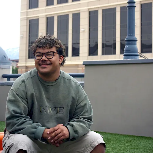 A young man with curly hair and glasses sits on grass near a modern beige building and lamp post, wearing a green sweatshirt and shorts, smiling.