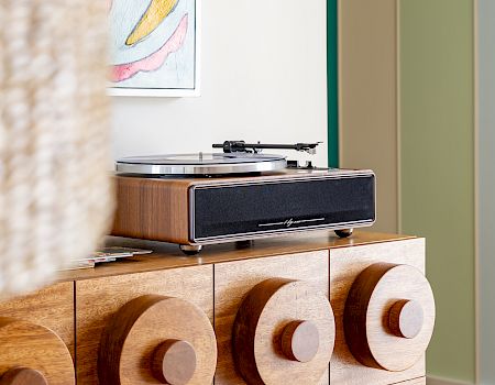 Un tocadiscos de madera reposa sobre un mueble decorativo, con coloridos cuadros colgados encima.