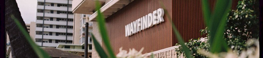 A city building entrance with a &ldquo;WAYFINDER&rdquo; sign, framed by green plants and tall high-rise apartments in the background.