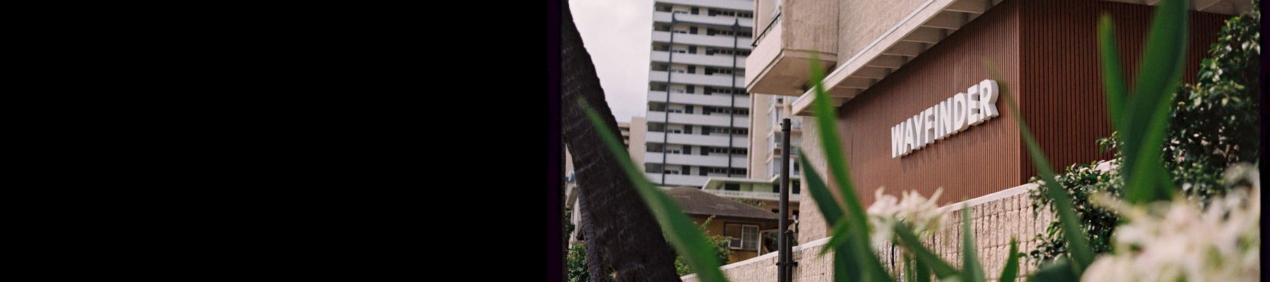A city building entrance with a &ldquo;WAYFINDER&rdquo; sign, framed by green plants and tall high-rise apartments in the background.