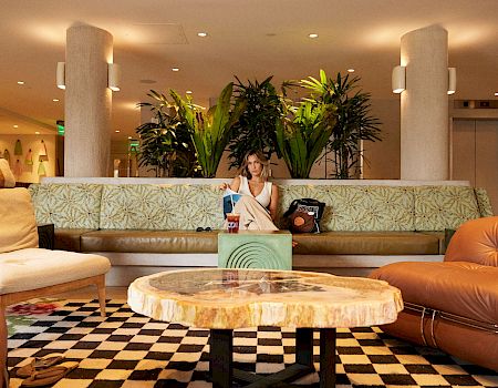 Una mujer descansa en un sofá estampado en el vestíbulo de un hotel, rodeada de grandes plantas en macetas y una iluminación cálida.