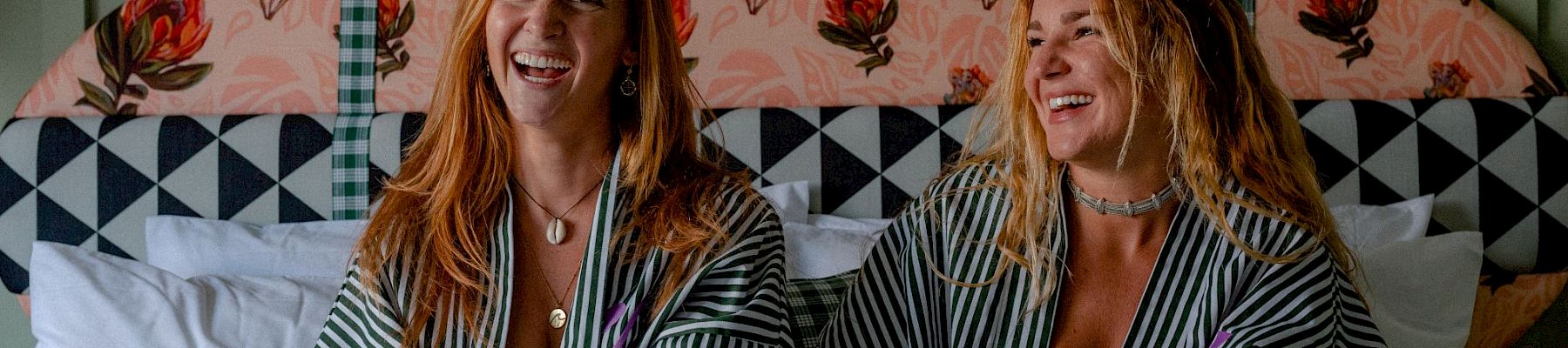 Two women in striped pajamas laugh together on a bed, relaxing with floral and checkered bedding behind them.