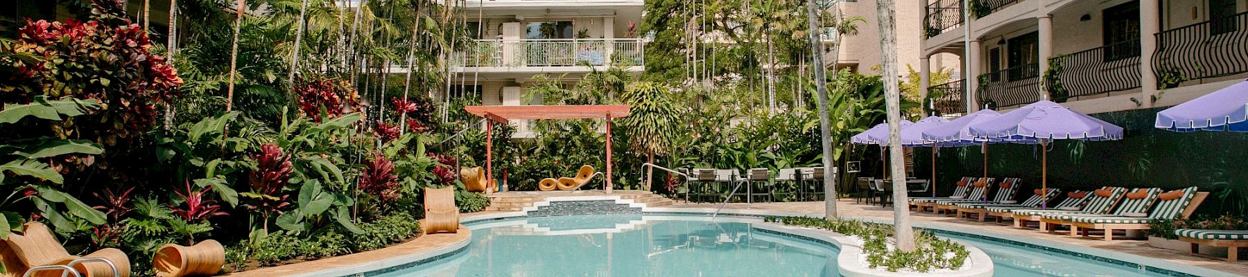A sunny hotel courtyard with a large outdoor swimming pool, palm trees, lush gardens, and lounge chairs under purple umbrellas.