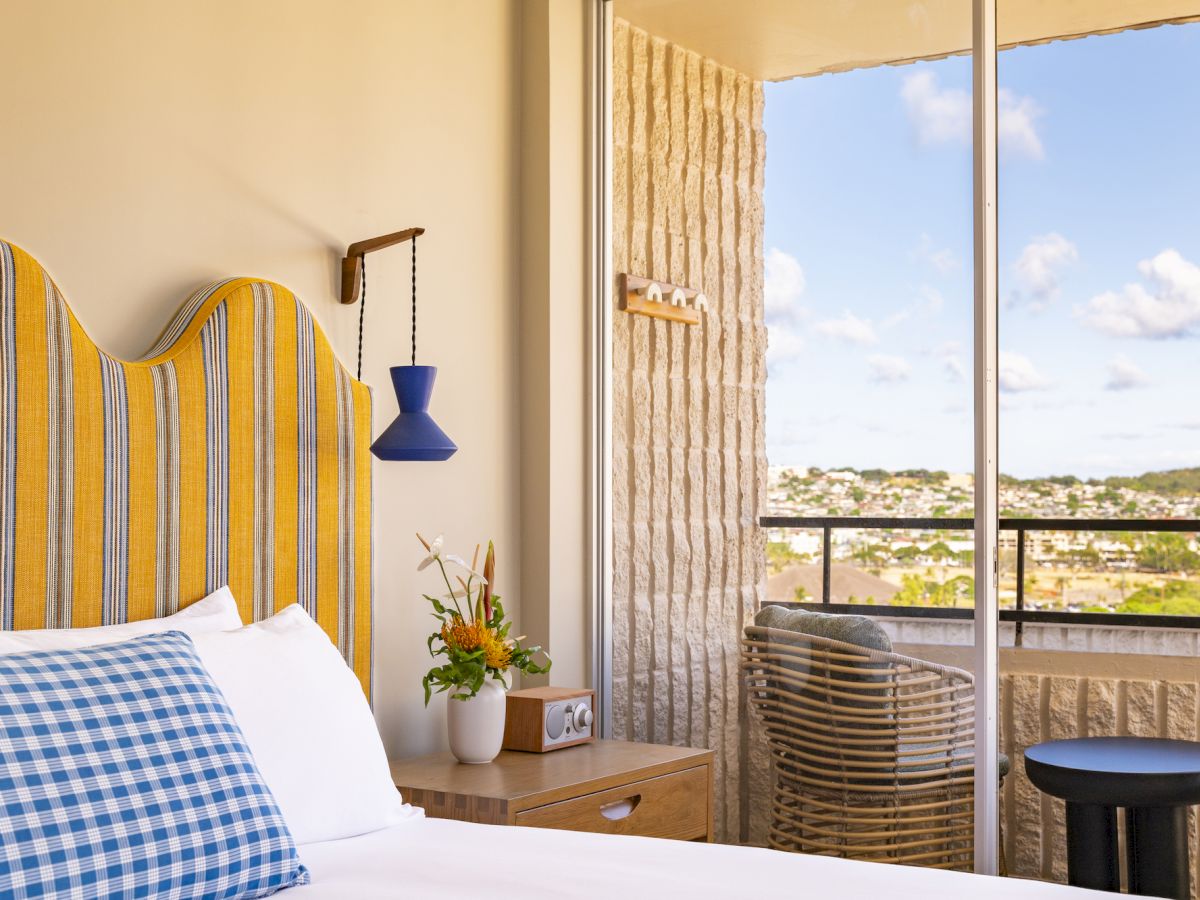 A bright bedroom with striped headboard, blue pendant lamp, flowers on the nightstand, and an open balcony view.
