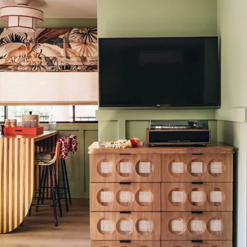 Lichtdurchflutete Wohnk&uuml;che mit Wandfernseher, gemustertem Rollo, Holzbar, Schrank und gestreifter H&auml;ngesch&uuml;rze.