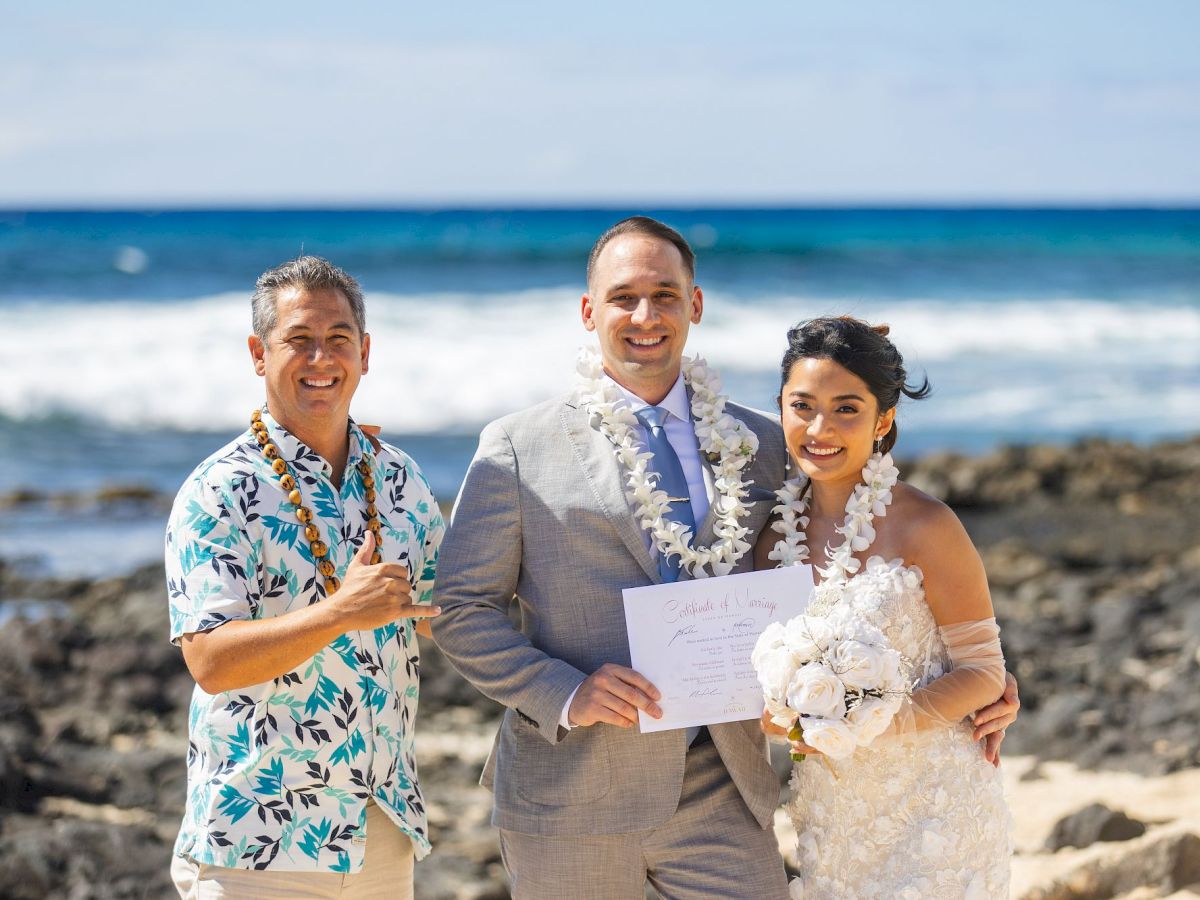 A groom, bride, and officiant pose on a sunny beach with leis and a signed marriage certificate in hand.