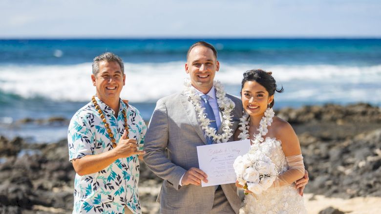 A groom, bride, and officiant pose on a sunny beach with leis and a signed marriage certificate in hand.