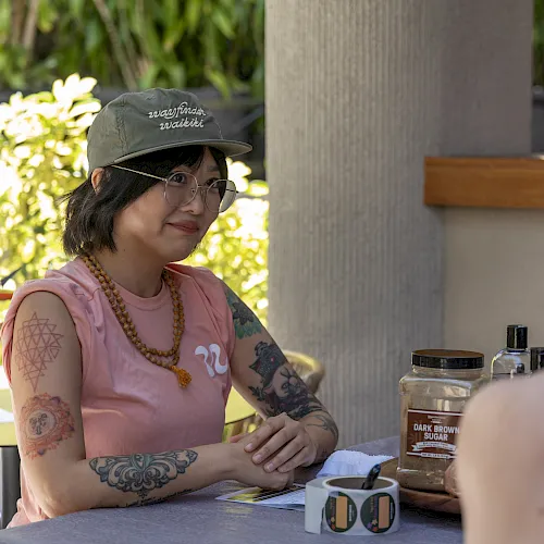 An outdoor caf&eacute;: a woman in a cap and glasses chats while a table holds spice/sugar jars and her tattoos show.