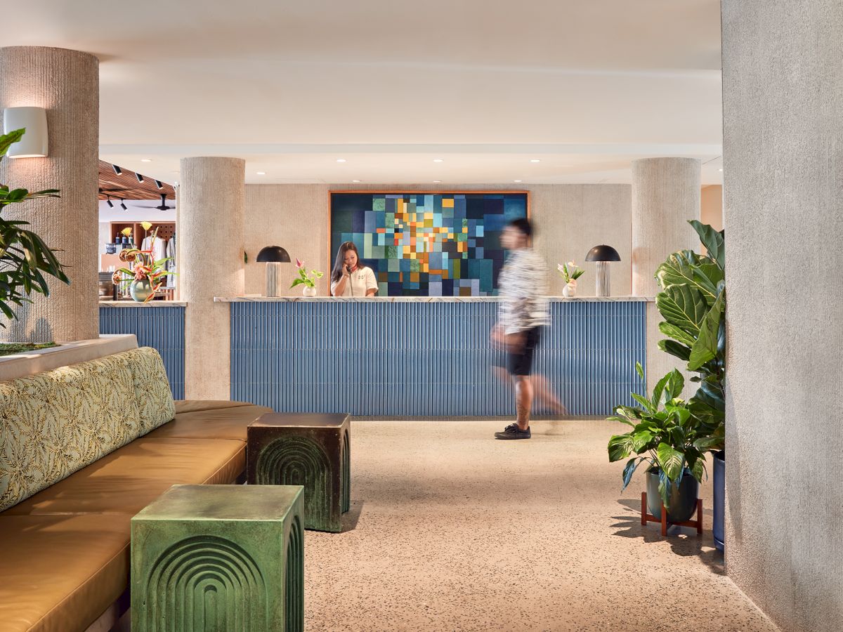 A modern hotel/lobby lounge with plants, cushioned seating, and a reception desk where people are gathered, under warm lights.