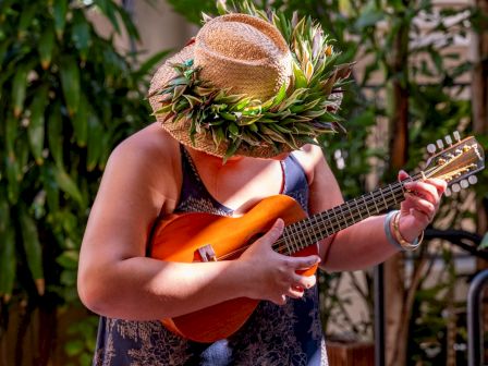 Un m&uacute;sico con un sombrero de paja floreado toca un ukelele naranja en un frondoso patio ajardinado.