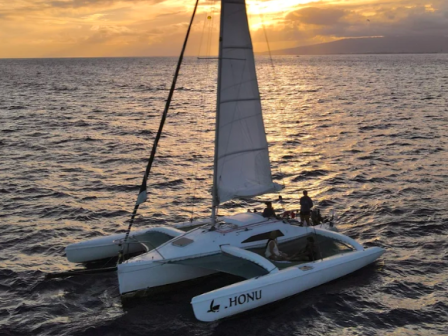 Un catamar&aacute;n de vela con la vela mayor izada flota a la deriva en el oc&eacute;ano al atardecer, con la palabra "HONU" en su casco.