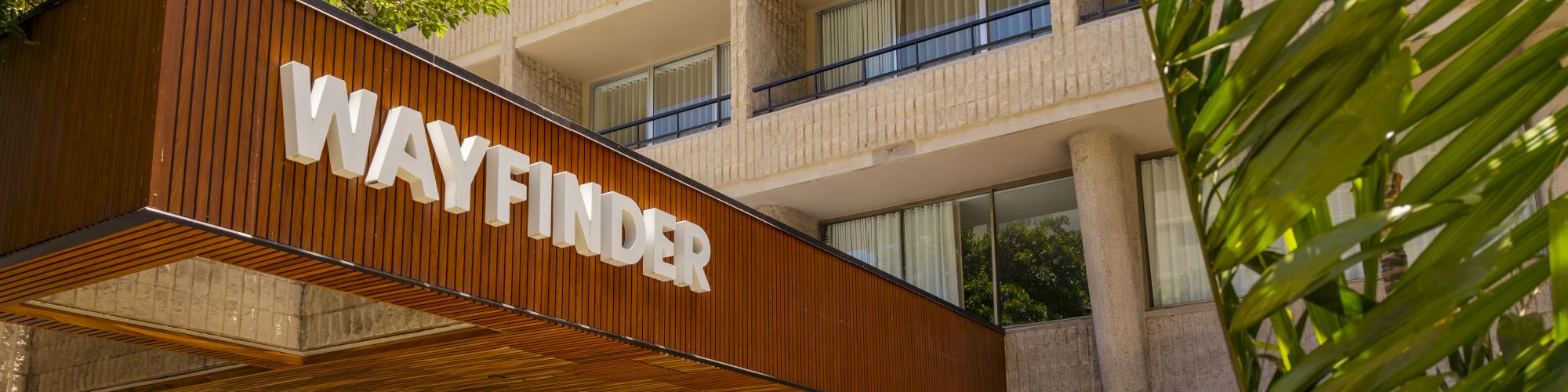 A sunny hotel courtyard with lush plants and a large &ldquo;WAYFINDER&rdquo; sign on a wooden wall under apartment balconies.