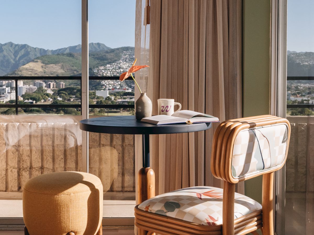 A sunny balcony reading nook with a small round table, chair, vase with flowers, an open book, and mountain views outside.