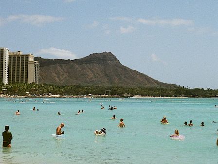 People float and swim in a bright turquoise ocean at a beach, with jagged mountains and hotel buildings in the background.