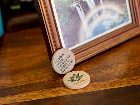 A wooden table shows two round green-labeled coasters beside a tilted photo frame with a mountain waterfall view.