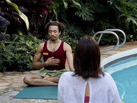 A man in a maroon tank leads meditation/yoga by a pool while a woman faces him in a lush garden.