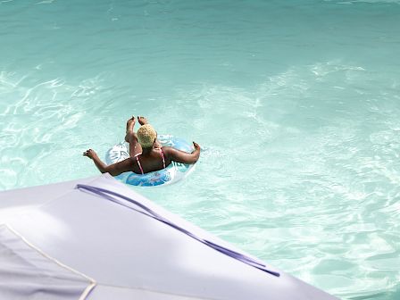 A swimmer lounges on an inner-tube in a turquoise pool, surrounded by lush tropical plants and palm leaves in the sun.